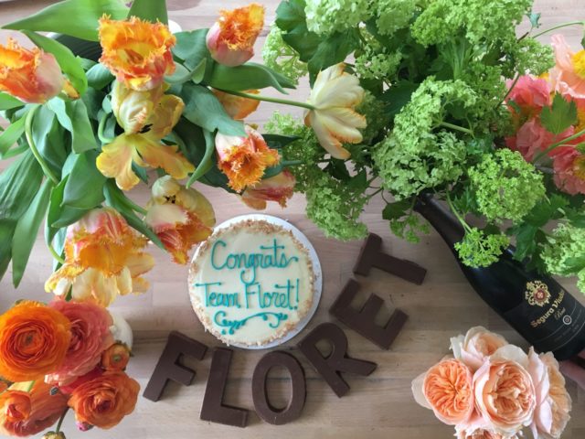 A cake that reads "congrats team floret!" and chocolate letters spelling "floret" on a table surrounded by flowers