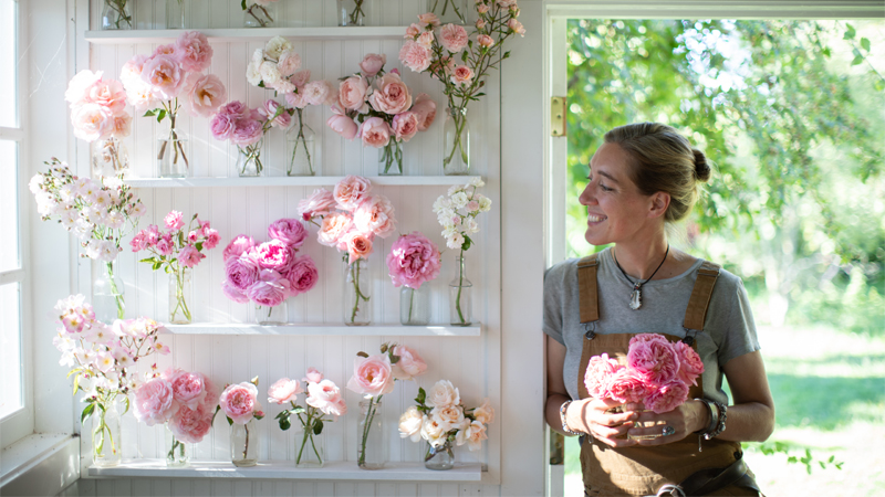Erin Benzakein smiling and looking at pink-toned rose blooms lining the wall of her studio