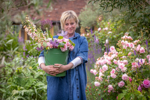 Sarah Raven carrying bucket of June flowers including foxgloves, roses, clematis, geraniums and later flowering alliums